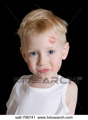 Stock Image - A young blond haired blue-eyed boy wearing white vest with red lipstick kisses on his face. Studio lighting, black background.. Fotosearch