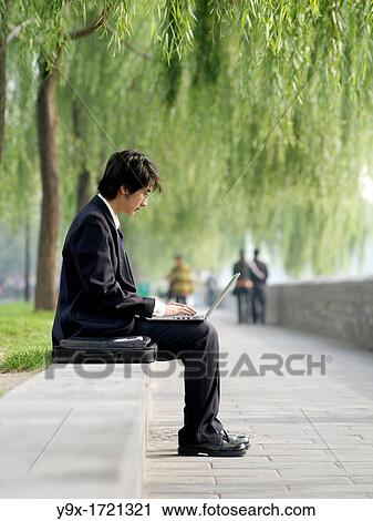 A young Chinese business man in a suit checking his email on his laptop through wireless internet in a nature park View Large Photo Image Stock Image - A young Chinese business man in a suit checking his email on his laptop through wireless internet in a nature park. Fotosearch