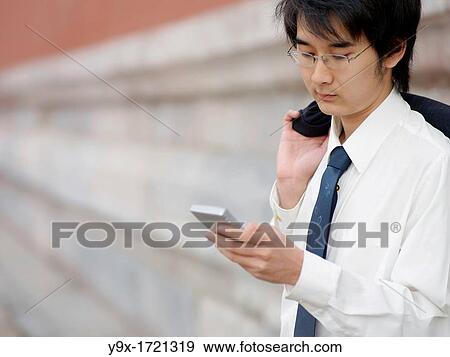 A young Chinese businessman in a shirt with a suit over his shoulder checking his text messages or email on his mobile smart phone through wireless internet with traditional Chinese architecure in the background in Beijing View Large Photo Image Stock Photo - A young Chinese businessman in a shirt with a suit over his shoulder checking his text messages or email on his mobile smart phone through wireless internet with traditional Chinese architecure in the background in Beijing. Fotosearch