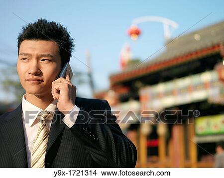 A young Chinese businessman in a suit checking his text messages or email on his mobile smart phone through wireless internet with traditional Chinese architecure in the background in Beijing View Large Photo Image Picture - A young Chinese businessman in a suit checking his text messages or email on his mobile smart phone through wireless internet with traditional Chinese architecure in the background in Beijing. Fotosearch