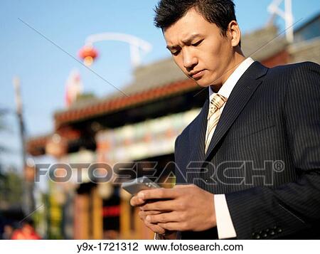 Stock Image - A young Chinese businessman in a suit checking his text messages or email on his mobile smart phone through wireless internet with traditional Chinese architecure in the background in Beijing. Fotosearch