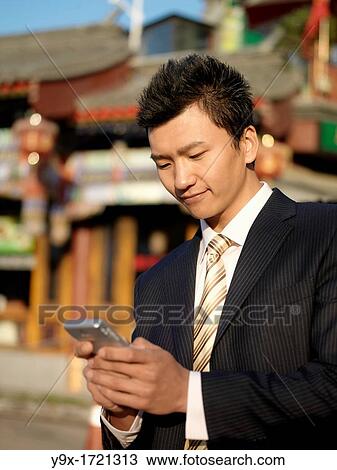 Stock Image - A young Chinese businessman in a suit checking his text messages or email on his mobile smart phone through wireless internet with traditional Chinese architecure in the background in Beijing. Fotosearch