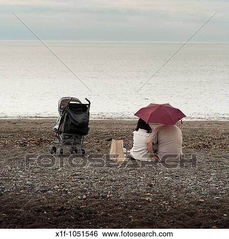 A young couple sitting under an umbrella on a deserted beach on a typically british wet summer afternoon with a child's buggy next to them Wales UK View Large Photo Image Stock Photograph - A young couple sitting under an umbrella on a deserted beach on a typically british wet summer afternoon with a child's buggy next to them Wales UK. Fotosearch