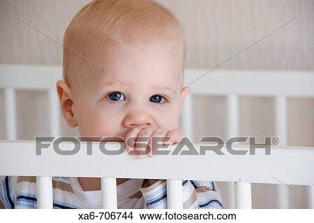 Picture - A young fair-haired, blue-eyed baby boy stands in his white crib, looking at the camera.. Fotosearch
