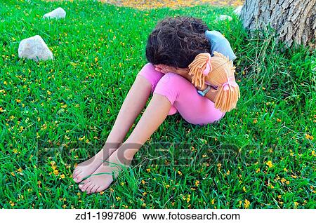 Stock Photograph - A young girl hides her face and cries under a tree on the grass while holding her stuffed toy doll.. Fotosearch
