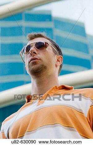 Stock Image - A young man in casual dress wearing sunglasses stands in front of a modern high rise building. Fotosearch