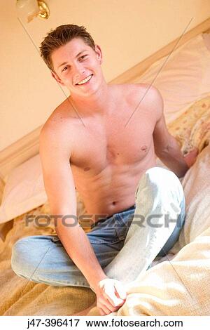 A young man sitting on a bed looking into camera smiling View Large Photo Image Stock Photo - A young man sitting on a bed looking into camera smiling. Fotosearch