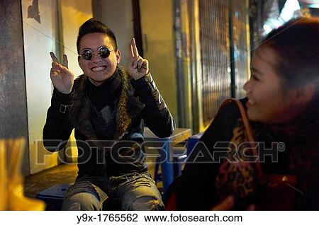 A young man with sunglasses poses with victory signs at an outdoor restaurant while a young woman looks on View Large Photo Image Stock Image - A young man with sunglasses poses with victory signs at an outdoor restaurant while a young woman looks on. Fotosearch
