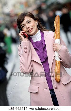 Picture - A young woman holding a baguette and talking on her cell phone on the street of Paris. Paris. France.. Fotosearch