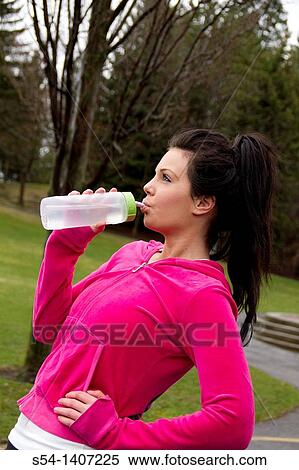 Stock Photography - A young woman in workout clothes at the park, drinking water. Fotosearch