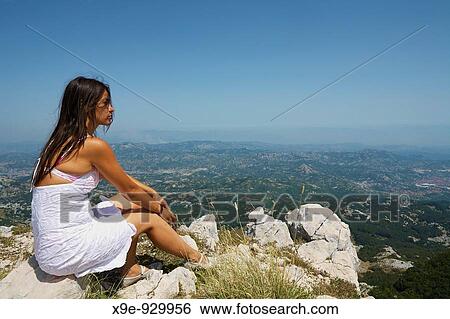 Stock Photograph - A young woman is sitting at the top of the rock looking ahead. Fotosearch