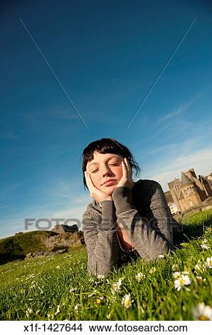 Picture - A young woman lying on the grass outdoors with her head in her hands daydreaming on a spring summer afternoon UK. Fotosearch