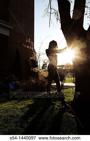 Stock Photo - A young woman standing by a tree, backlit by the sun.. Fotosearch