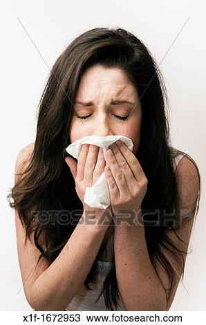 Stock Image - A young woman with brown hair and eyes feeling ill sneezing blowing her nose into a paper tissue. Fotosearch