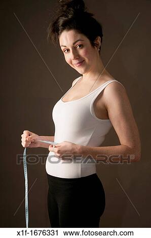 Stock Image - A young woman with brown hair and eyes measuring her body. Fotosearch