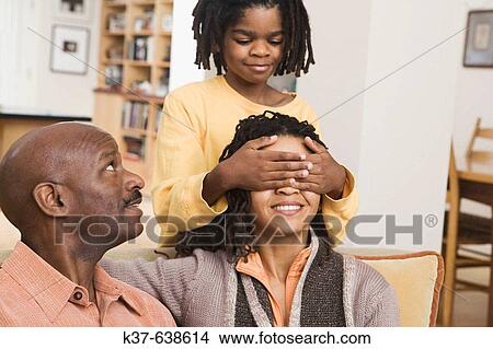 African American family at home, parents on sofa, son covering mother's eyes with hands from behind. Guess who? View Large Photo Image Picture - African American family at home, parents on sofa, son covering mother's eyes with hands from behind. Guess who?. Fotosearch