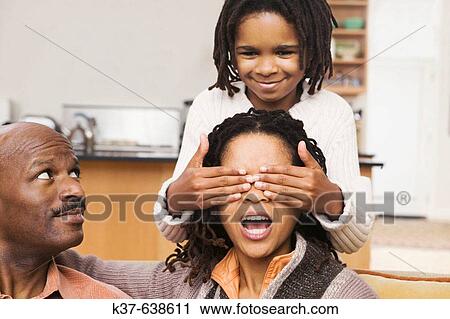 African-American family at home, parents on sofa & daughter covering mother's eyes with hands from behind. Guess who? View Large Photo Image Stock Image - African-American family at home, parents on sofa & daughter covering mother's eyes with hands from behind. Guess who?. Fotosearch