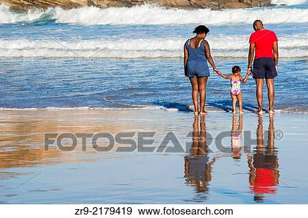 Stock Photo - African couple holding the hands of their todddler daughter as they approach waves at the surfing beach, Victoria Bay, Garden Route, Western Cape, South Africa.. Fotosearch