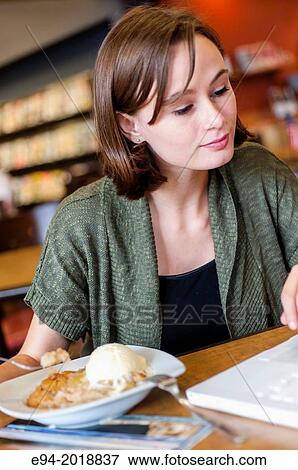 Stock Photo - An 18 year old girl in a library reading from a computer screen with a dessert on the table.. Fotosearch