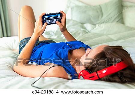 An 18 year old girl lying on her bed texting and weaing head phones. View Large Photo Image Picture - An 18 year old girl lying on her bed texting and weaing head phones.. Fotosearch