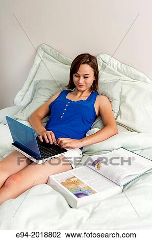 Stock Image - An 18 year old girl sitting on her bed with a computer, text book and cell phone.. Fotosearch