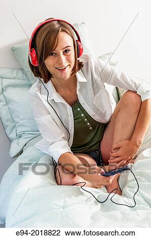 Stock Image - An 18 year old girl sitting cross-legged on her bed wearing head phones and holding a cell phone.. Fotosearch