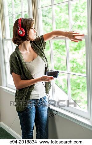 An 18 year old girl standing by a window in a house looking outside wearing head phones and holding a cell phone. View Large Photo Image Picture - An 18 year old girl standing by a window in a house looking outside wearing head phones and holding a cell phone.. Fotosearch