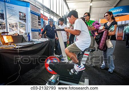 Stock Image - An Asian boy using a pedal powered generator works to create enough electricity to light a lamp at the Solar Decathlon in Irvine, CA. The international competition is sponsored by the US Department of Energy.. Fotosearch