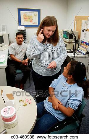 An audiologist uses sign language to communicate with a hearing impaired adolescent Hispanic girl at a speech and hearing clinic in Orange, California. Note new hearing aid components on table and parent at left View Large Photo Image Stock Photo - An audiologist uses sign language to communicate with a hearing impaired adolescent Hispanic girl at a speech and hearing clinic in Orange, California. Note new hearing aid components on table and parent at left. Fotosearch