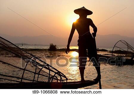 Stock Photo - An Intha fisherman with traditional fish trap uses an unusual leg-rowing technique to propel his flat-bottomed boat across the lake while standing.. Fotosearch