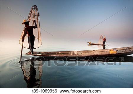 Stock Photograph - An Intha fisherman with traditional fish trap uses an unusual leg-rowing technique to propel his flat-bottomed boat across the lake while standing.. Fotosearch