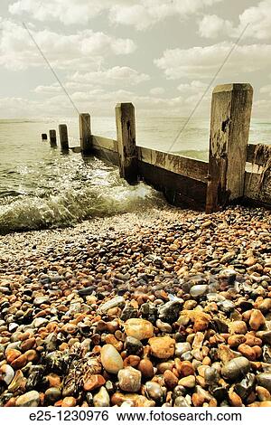 Stock Photograph - An old breakwater sea defence on a pebble beach with a small wave from a calm sea breaking up the beach. Location: Sussex UK. Fotosearch
