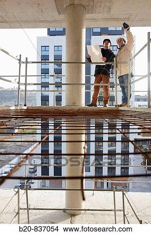 Picture - Architect and construction worker looking at blueprints, personal protective equipment, housing construction, concrete skeleton. Fotosearch