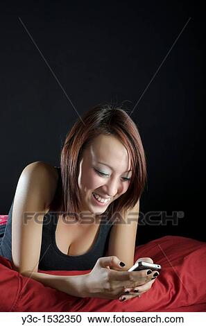 Stock Image - Asian girl in bed playing with iphone. Fotosearch