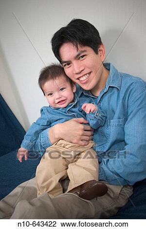 Stock Image - Asian man sitting on a bed with his Eurasian toddler son.. Fotosearch