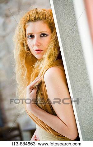Stock Image - Attractive young woman is leaning on a wall, looking over her shoulder and touching long curly hair. Fotosearch
