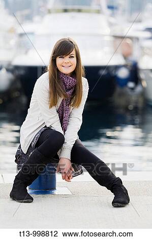 Stock Image - Attractive young woman is sitting on a small harbor bollard. Fotosearch
