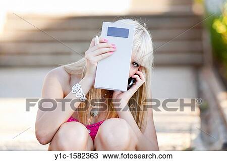 Stock Image - Attractive young woman is talking on a cellular phone and hiding behind a paper notebook. Fotosearch