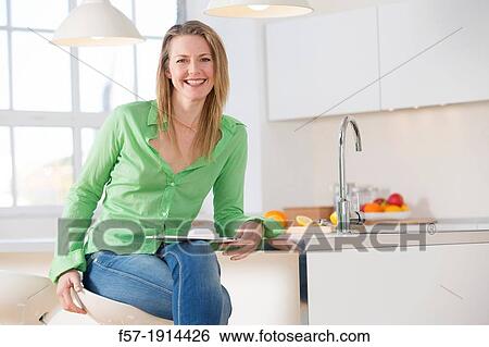 Stock Photograph - Attractive young woman using her tablet computer in her kitchen. Fotosearch