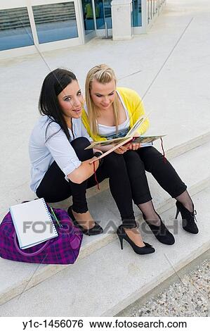 Stock Photograph - Attractive young women are sitting on low steps and looking at album full of photos. Fotosearch