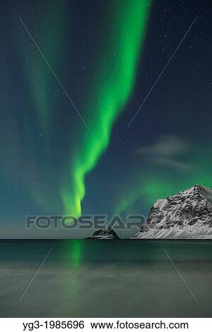 Stock Photograph - Aurora Borealis - Northern Lights shine in sky over snow covered mountains from Vik beach, Vestv? goy, Lofoten Islands, Norway. Fotosearch