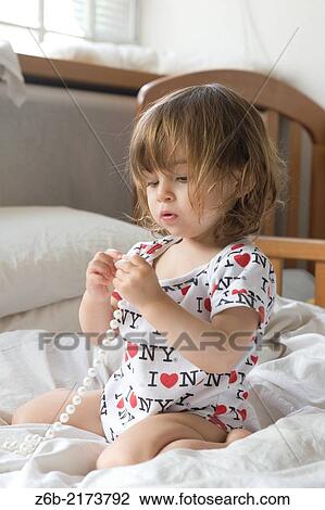 Stock Image - Baby girl, 20 months, Necklace, pearls, Bedroom, duvet, pillow, cot, Barefoot, Bodysuit, window. Fotosearch