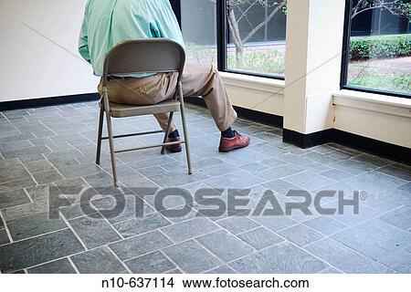 Picture - Back of a man sitting on a folding chair in an office building lobby. Fotosearch
