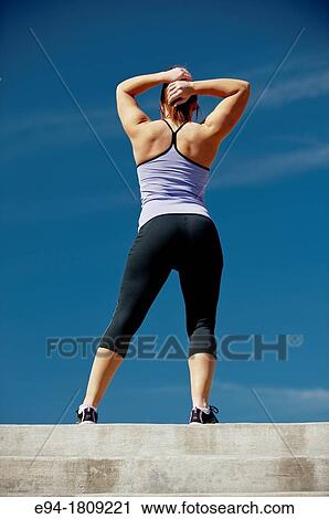 Stock Image - Back view of a 23 year old brunette woman in workout clothing outdoors against a blue sky. Fotosearch
