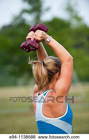 Back view of a woman in workout clothes holding a dumbbell above her head at a boot camp with other other women in the background. View Large Photo Image Stock Photo - Back view of a woman in workout clothes holding a dumbbell above her head at a boot camp with other other women in the background.. Fotosearch