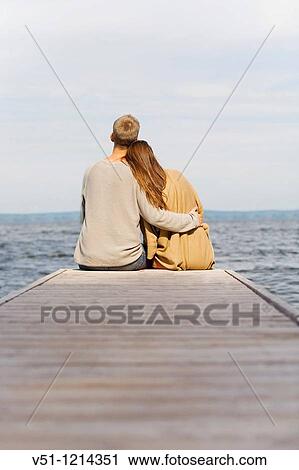 Stock Image - Back view of a young couple sitting in a pier. Fotosearch