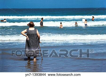 Back view of an old woman with her walking stick standing in the shallows at the beach watching younger people having fun in the waves View Large Photo Image Picture - Back view of an old woman with her walking stick standing in the shallows at the beach watching younger people having fun in the waves. Fotosearch