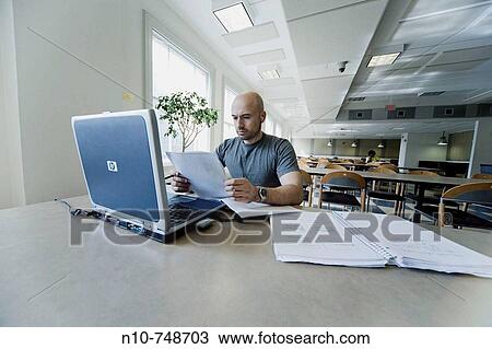 Stock Image - Balding young man working on his laptop computer in the dining area at his college.. Fotosearch
