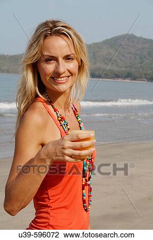 Stock Image - Beautiful blonde woman wearing an orange vest and drinking a glass of orange juice on a beach in India.. Fotosearch