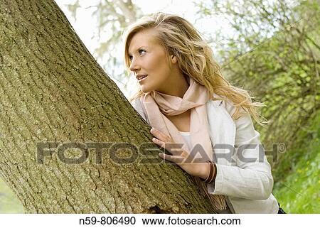 Stock Image - Beautiful young woman enjoying being outside in a green natural environment She is touching a tree in a contemporary twist on the idea of tree huggers. Fotosearch